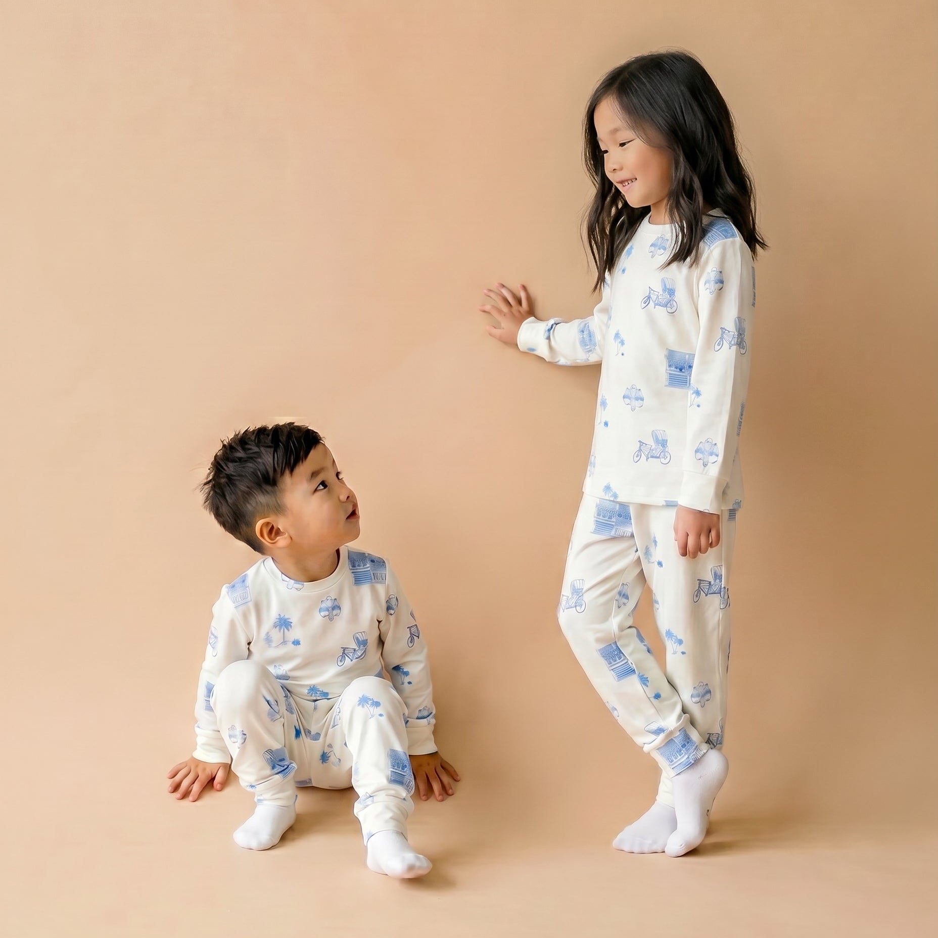 Two children wearing matching long-sleeve pajamas with a blue and white illustrated pattern, posed against a plain warm beige studio backdrop. One child stands and the other sits, both wearing white socks.