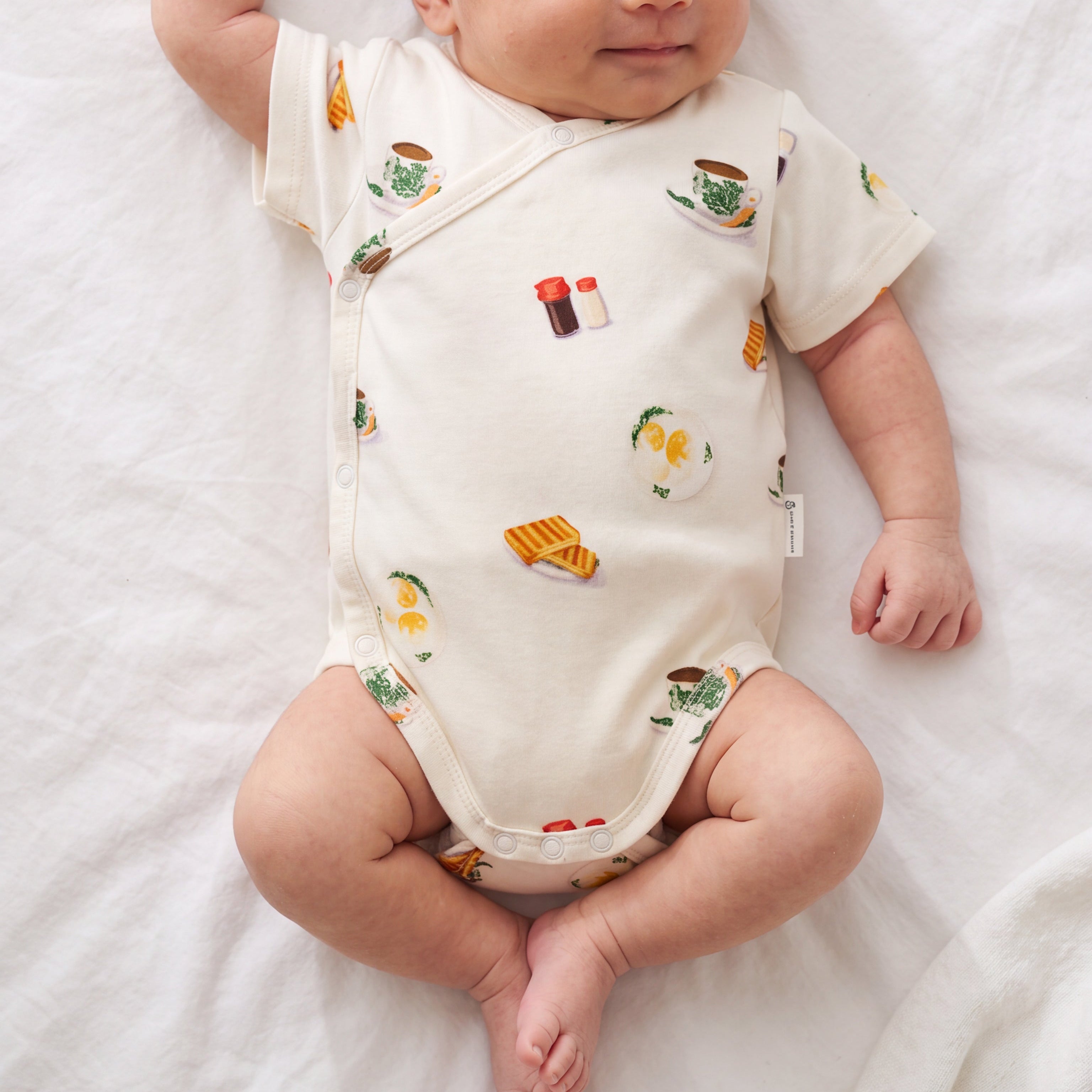 A sleeping newborn baby wearing a cream-colored short-sleeve bodysuit with colorful food illustrations, lying on a white fabric surface.